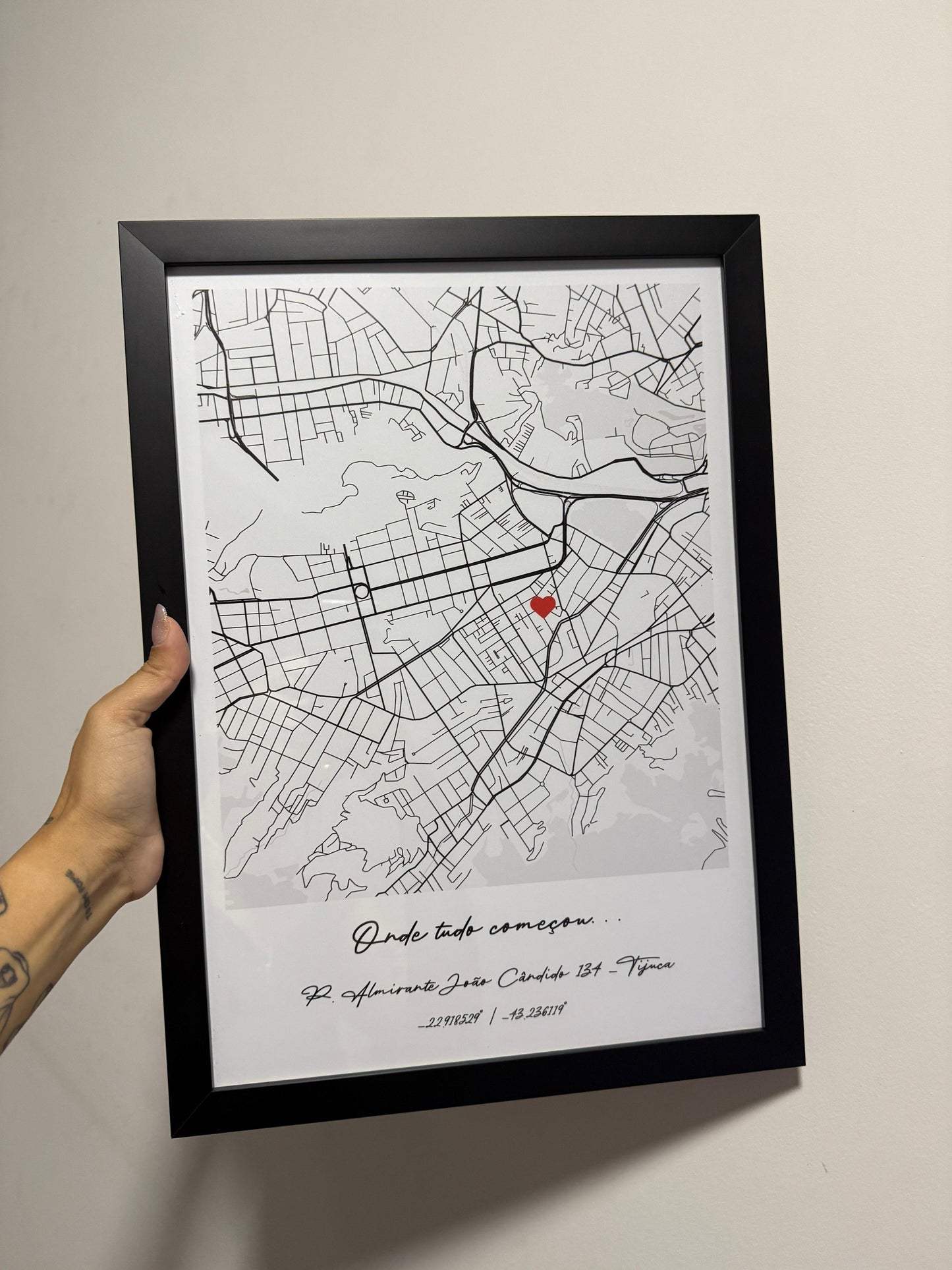 Mão segurando um quadro físico com moldura preta da Artmaps, mostrando um mapa personalizado com a frase 'Onde tudo começou' e um pin de coração vermelho.