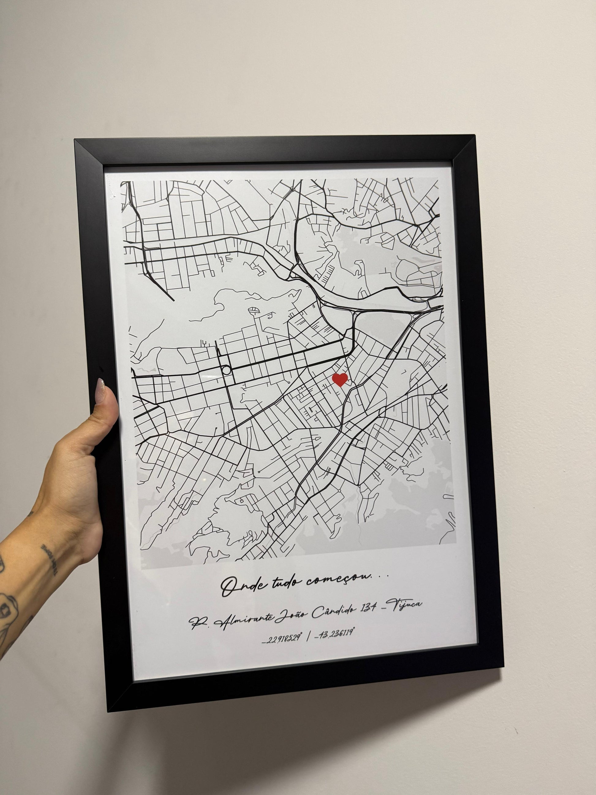 Mão segurando um quadro físico com moldura preta da Artmaps, mostrando um mapa personalizado com a frase 'Onde tudo começou' e um pin de coração vermelho.