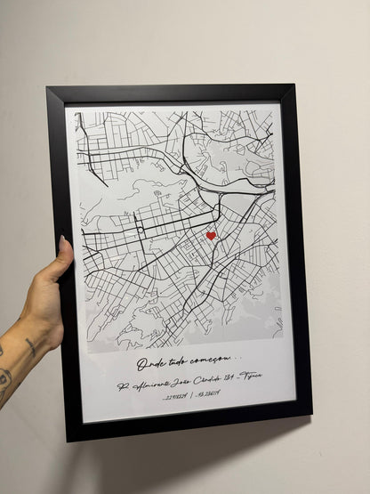 Mão segurando um quadro físico com moldura preta da Artmaps, mostrando um mapa personalizado com a frase 'Onde tudo começou' e um pin de coração vermelho.